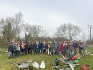 Around 40 people in outdoor clothes stood in a wide semi-circle, some holding garden spades and other tools in the air. Wheelbarrows and other garden tools are in the foreground